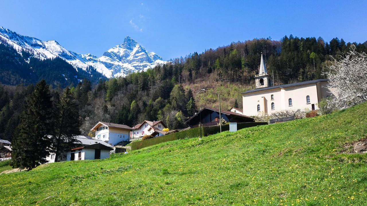 L’image est composée de trois plans en diagonale qui montent de gauche à droite. Au premier plan, on voit l’herbe verte d’un pâturage. Au-dessus, on aperçoit quelques chalets et une église aux murs blancs ornée de son clocher. Plus loin, derrière, on devine une colline couverte de sapins. En arrière-plan, des sommets enneigés se découpent sur un ciel uniformément bleu.