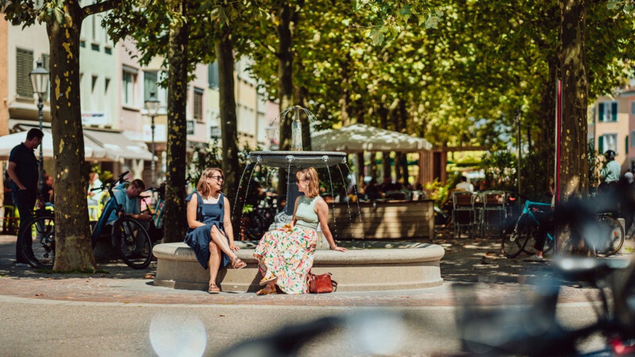 Two young women sitting together by a city fountain. 