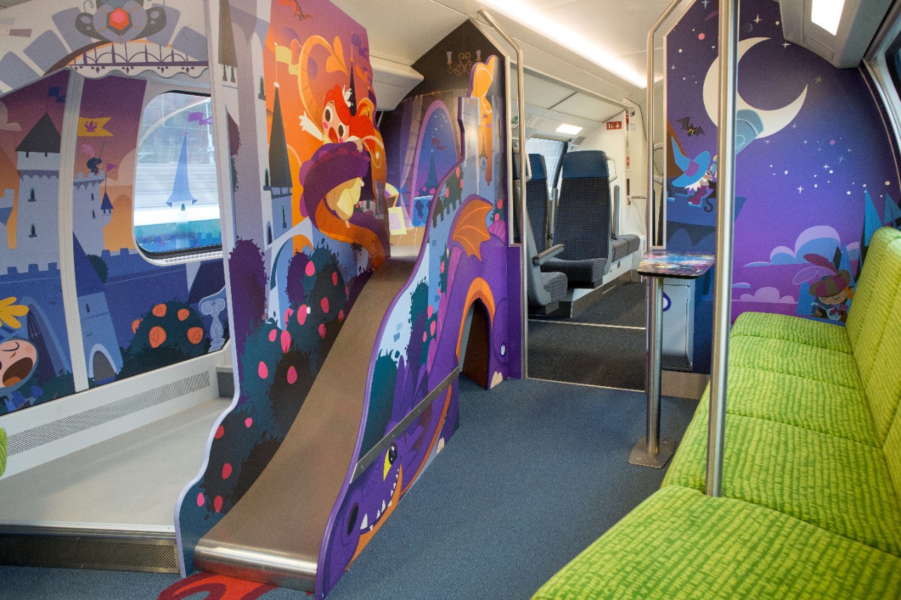 Carrozza famiglia sull’IC 200 – Al centro della carrozza si trova un piccolo scivolo su cui è raffigurato un drago. Sulla destra si trova una fila di sedili color verde chiaro.