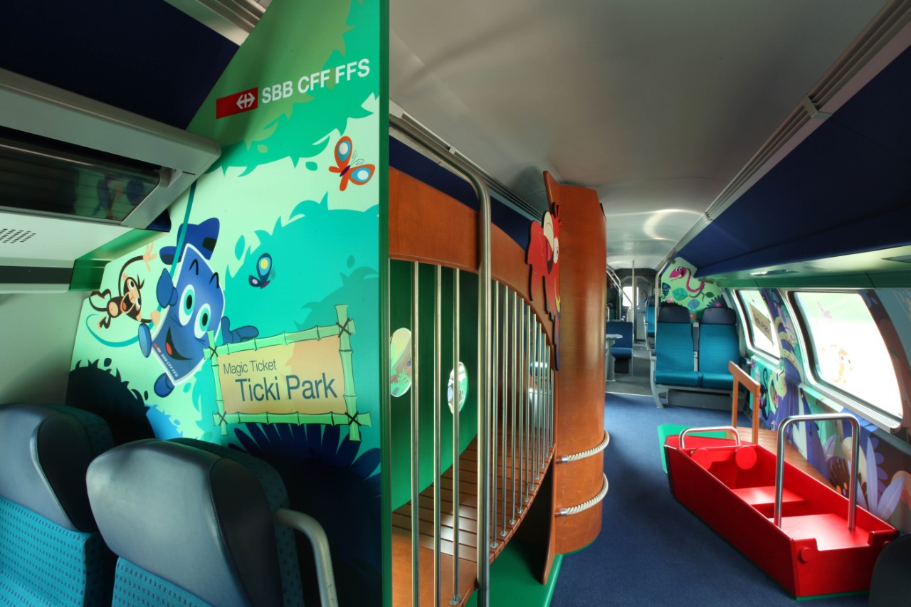 IC2000: Innenansicht des Ticki-Park-Familienwagens. Rechts im Bild ein rotes Holzschiff mit Haltestangen und sitzmöglichkeit. Daneben an der Wand ein „Steg“ um auf das Schiff zu gelangen. Im Fokus der Übergang zur Rutsche. Der Übergang ist links mit einer Wand und rechts mit Stangen gesichert, dass kein Kind hinunter fällt. Die Wände sind passend zum Thema „Dschungel“ gestaltet.