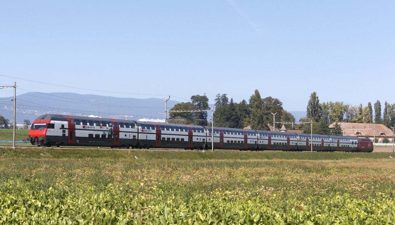 IC2000: Aussenansicht des IC2000. Der Zug, bestehend aus 6 Wagen und einer Lokomotive, fährt bei blauem Himmel durch die Landschaft. Im Hintergrund befinden sich Häuser, Bäume und Berge.