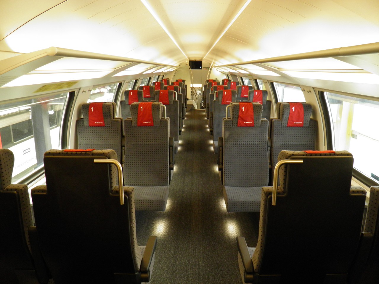 IR-Dost: Innenansicht der ersten Klasse. Hintem im Bild die Treppe in den unteren Bereich des Wagens. Oberhalb der Treppe befindet sich ein Bildschirm mit Kundeninformation. Unter den Sitzen, sowie oben in der Gepäckablage ist eine zusätzliche Beleuchtung eingebaut, dadurch heller. Am Boden ist Teppich verlegt.